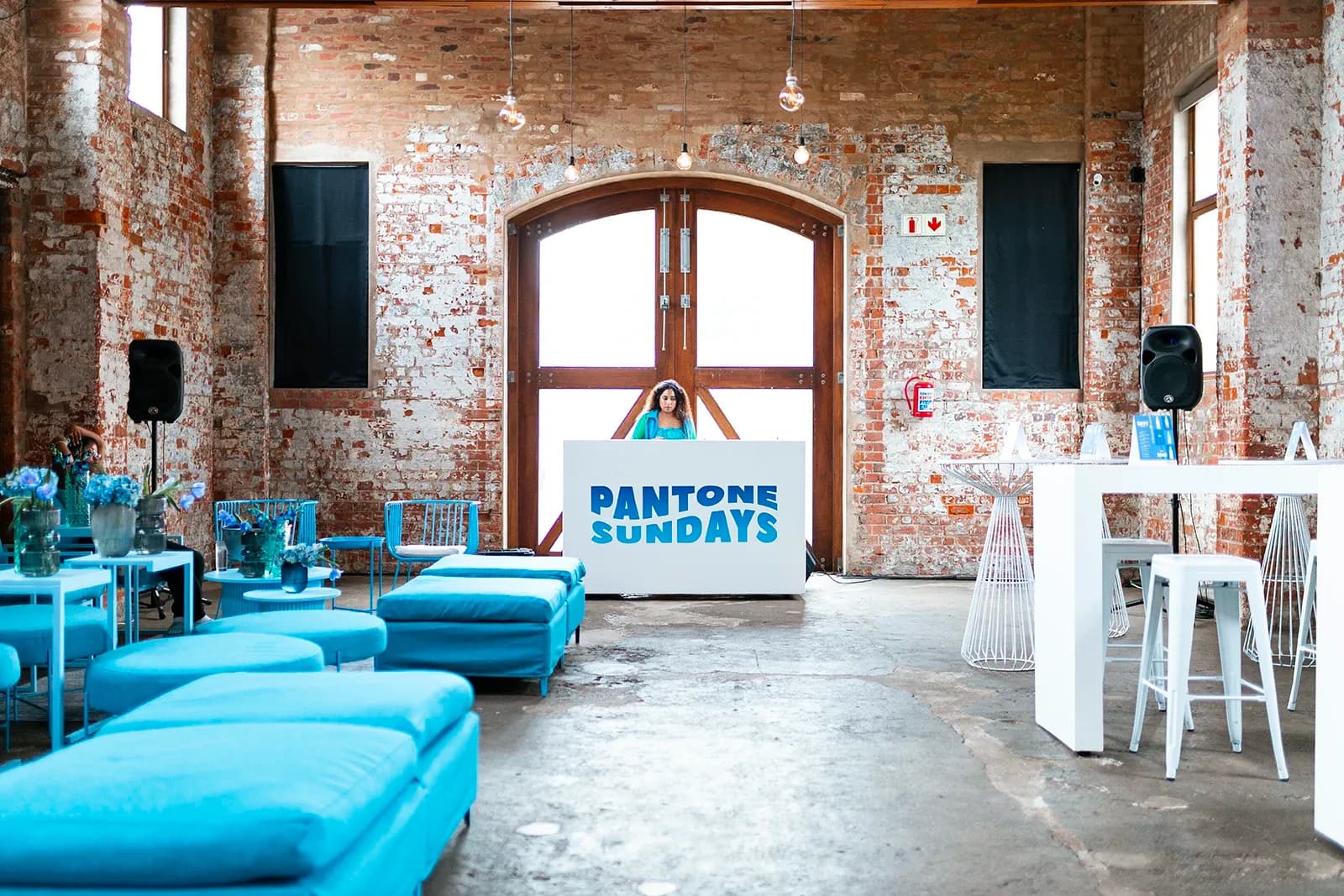 Interior event space featuring blue lounge furniture, exposed brick walls, and a DJ at a Pantone Sundays booth.