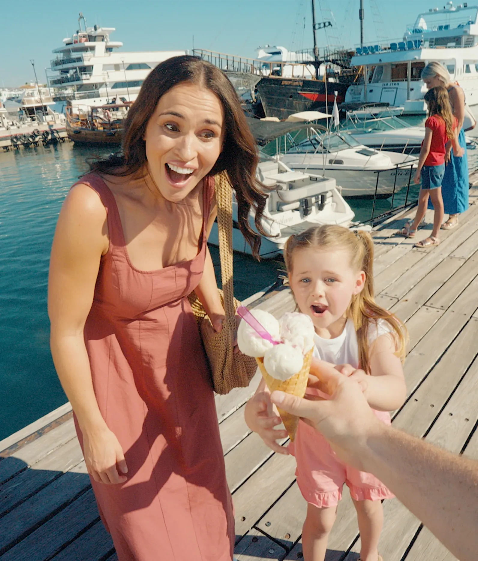POV shot handing an ice cream cone to a smiling woman and child on a sunny boat harbour dock.