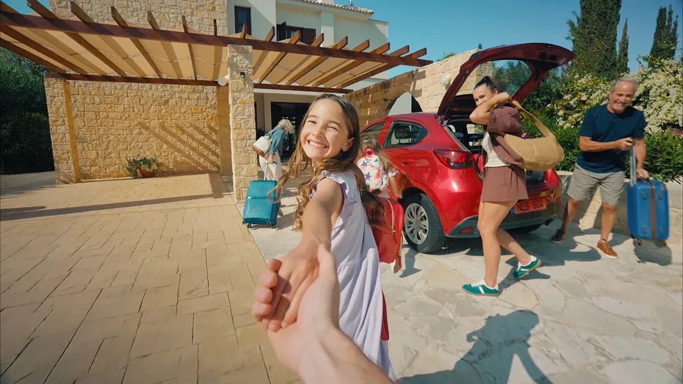 POV shot of a child leading someone toward a red rental car and stone villa while family members unload luggage.