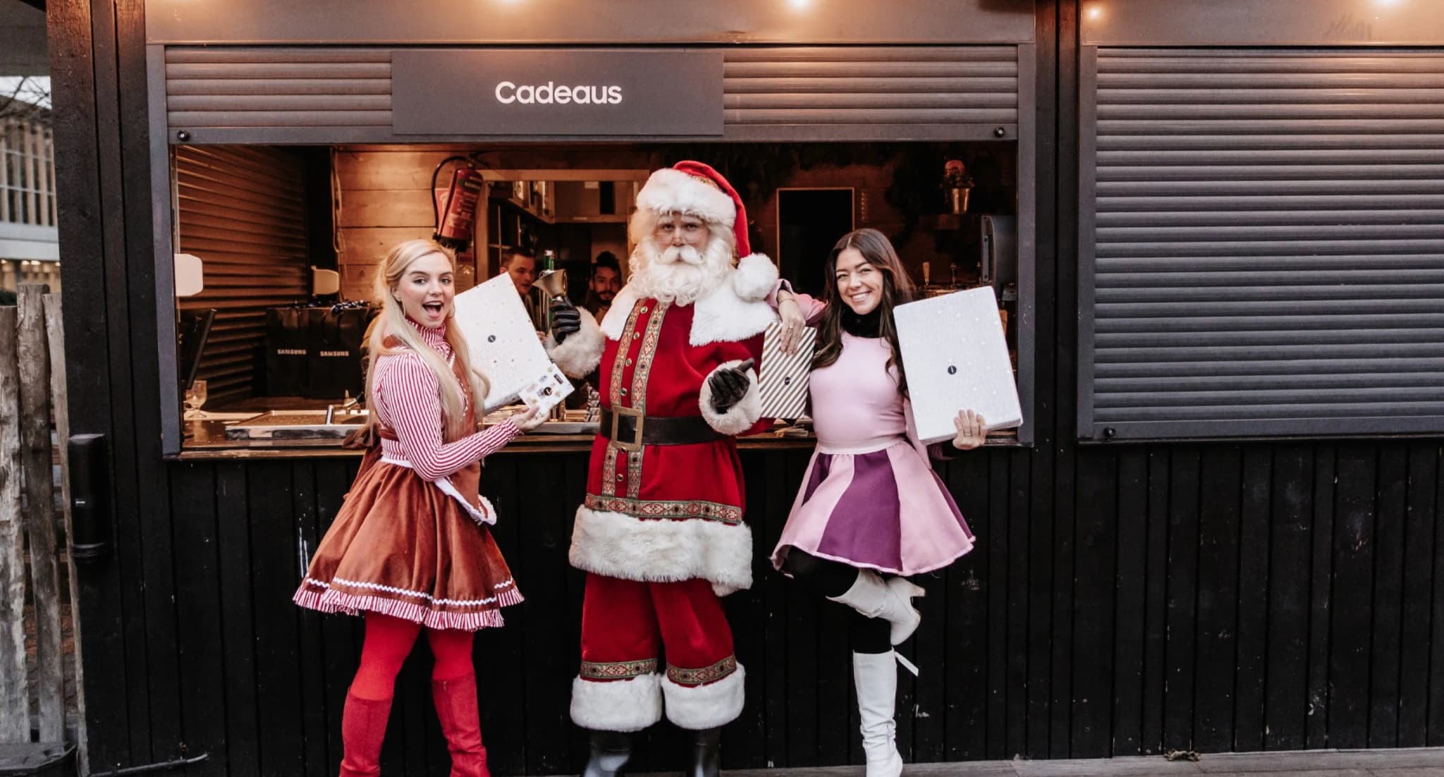 Santa Claus and two assistants holding gifts at a "Cadeaus" booth during a Samsung holiday event.