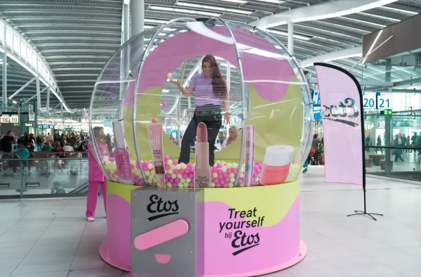 Woman posing inside a giant clear Etos ball pit display filled with pink and yellow balls and beauty product props.