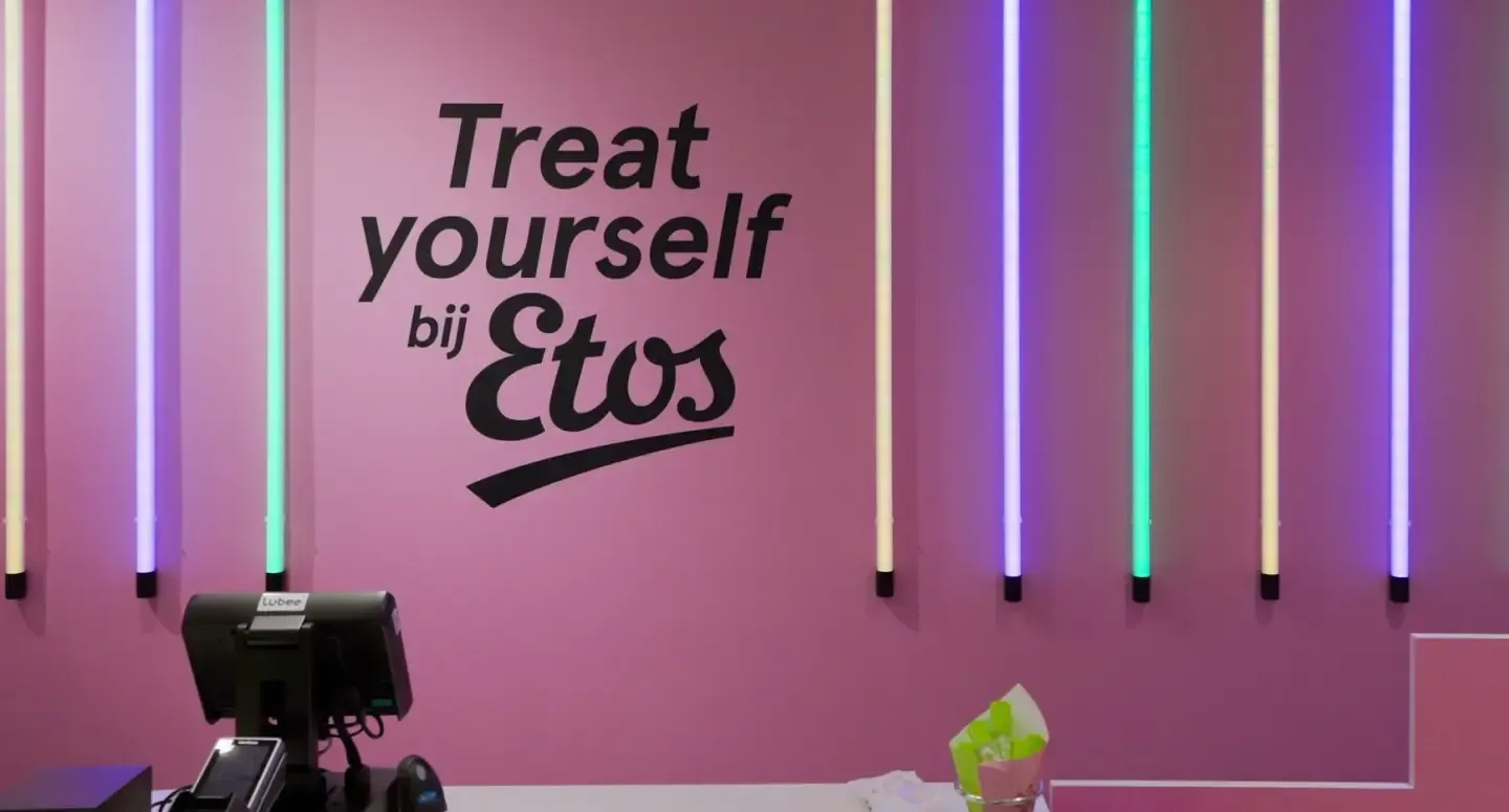 Pink wall featuring vertical neon lights and large black text reading "Treat yourself bij Etos" behind a service counter.