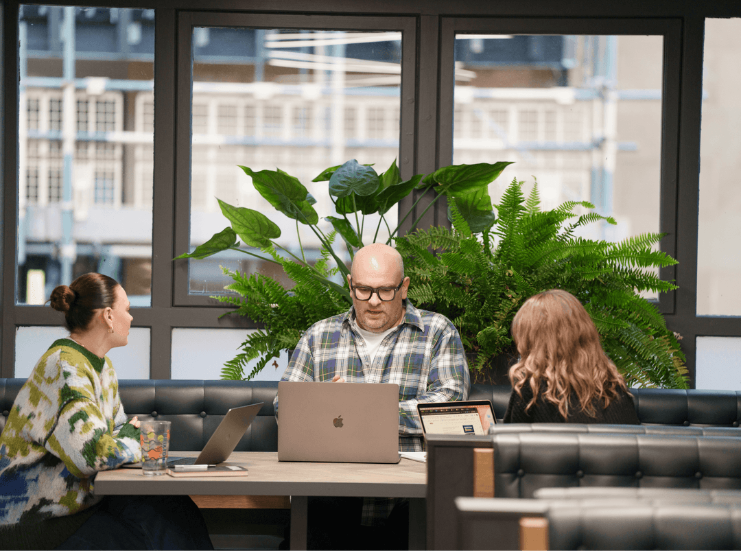 Colleagues sitting around a wooden table in a bright, modern London office with lush indoor plants.