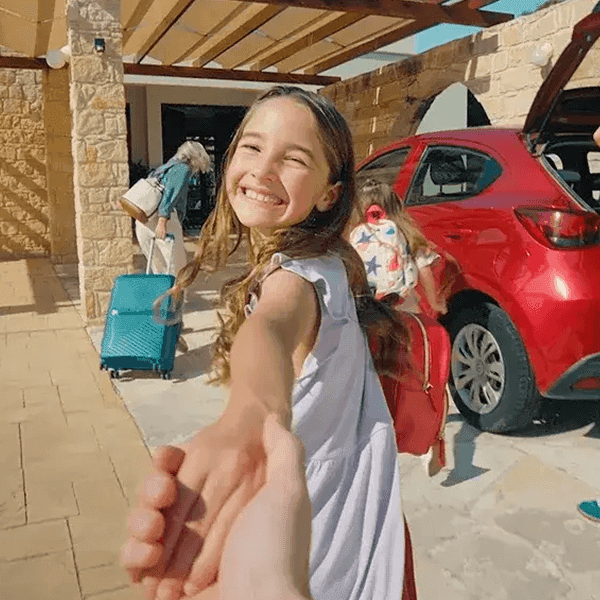 A smiling young girl holding a hand and leading someone toward a red car packed with luggage.