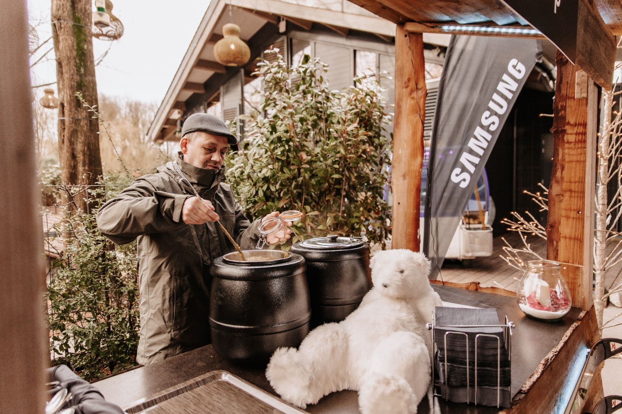 Man serving hot food at a winter market stall featuring a plush polar bear and Samsung branding.