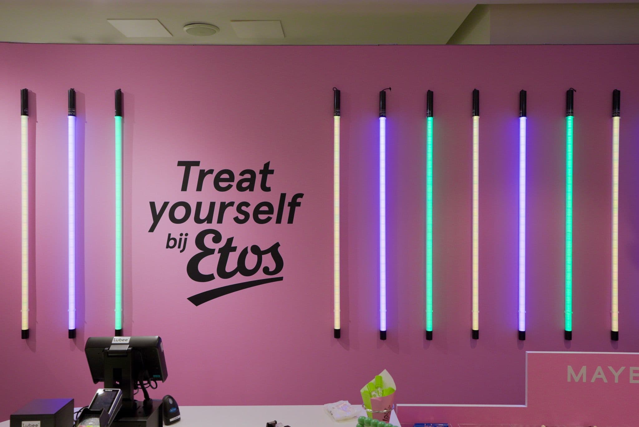 Pink wall featuring vertical neon lights and large black text reading "Treat yourself bij Etos" behind a service counter.