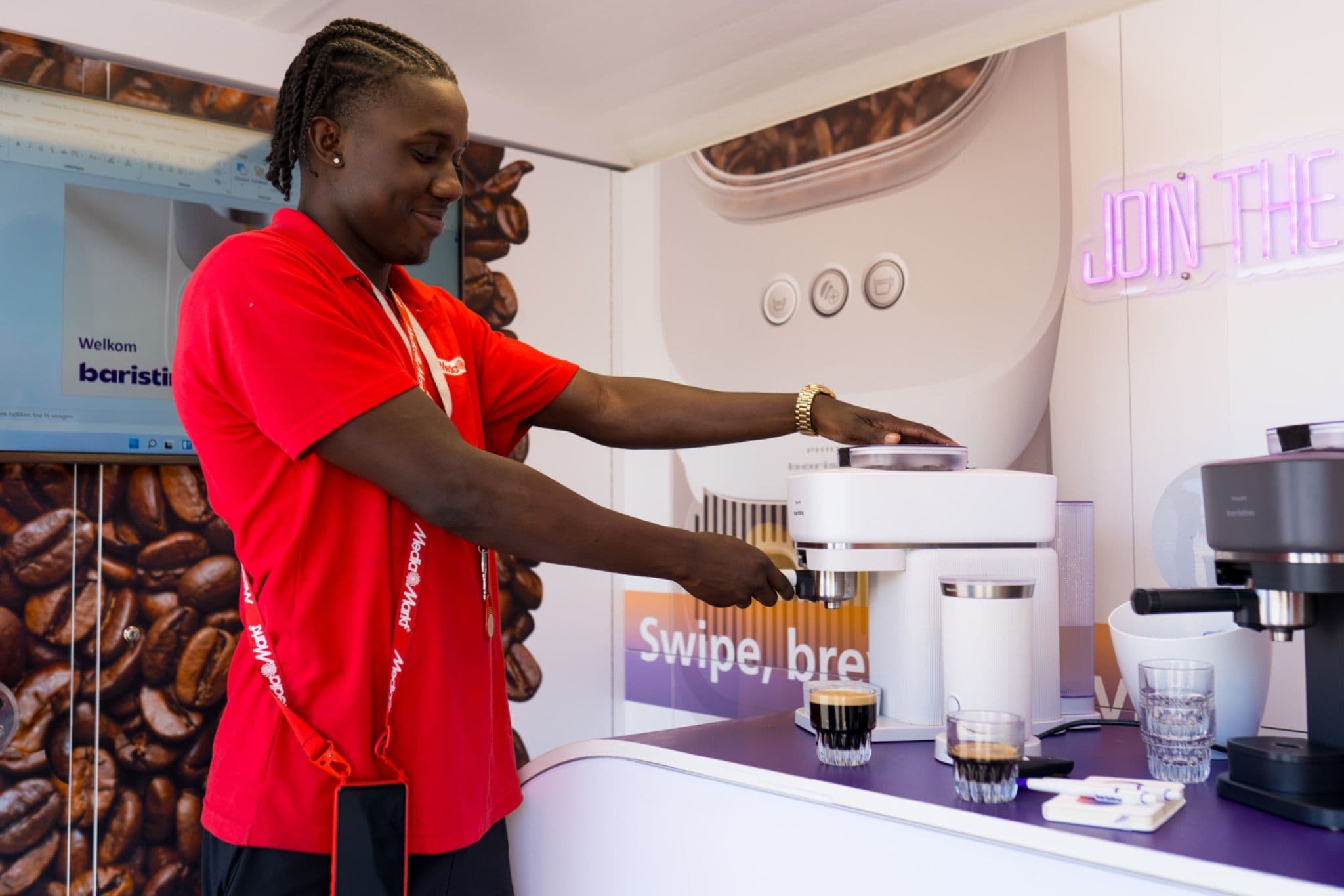 Staff member in a red shirt demonstrating a white Philips Baristina espresso machine at a promotional stand.