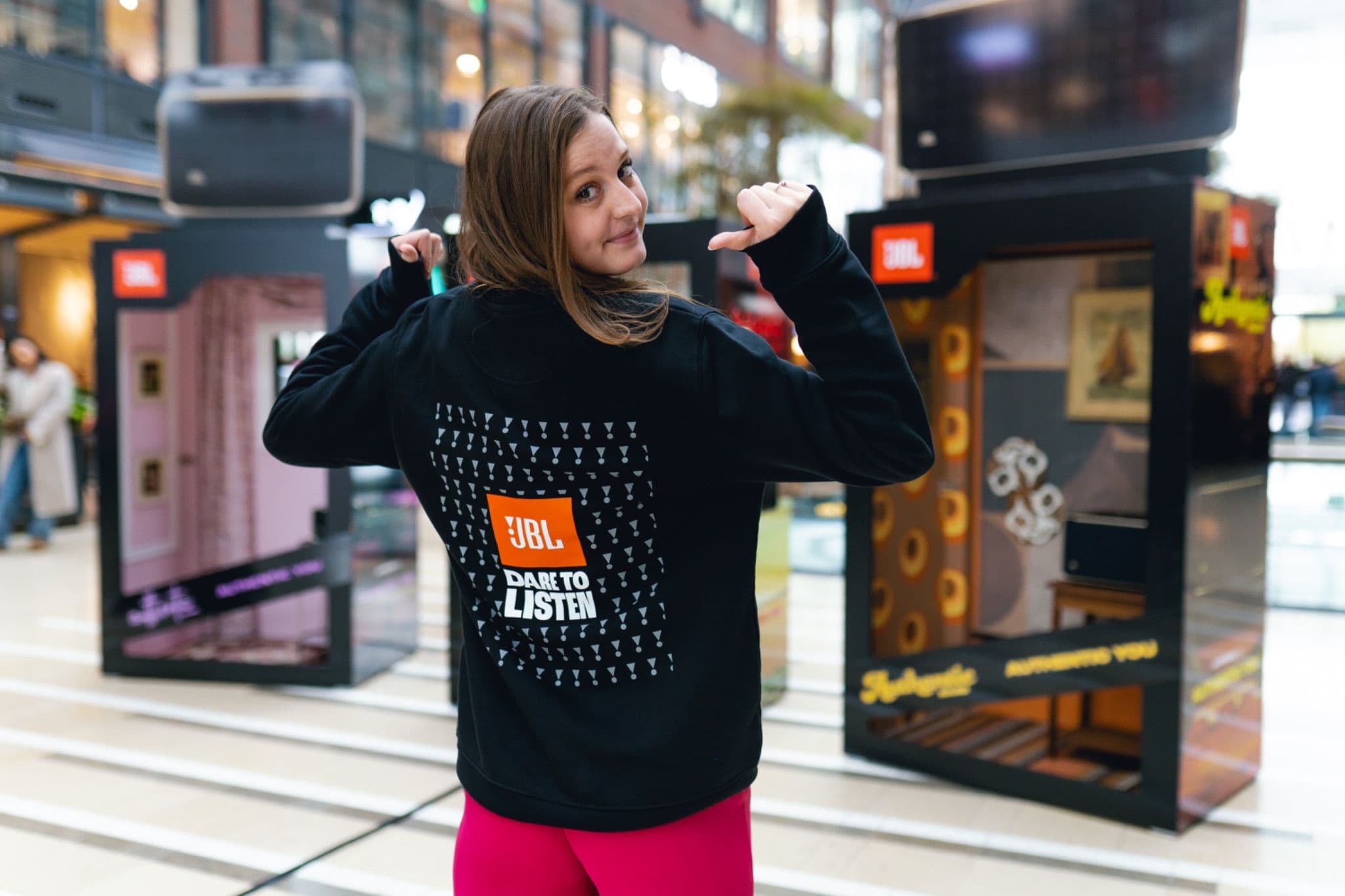 Woman wearing a black JBL hoodie with "Dare to Listen" slogan on the back, pointing to the logo at a promotional event.