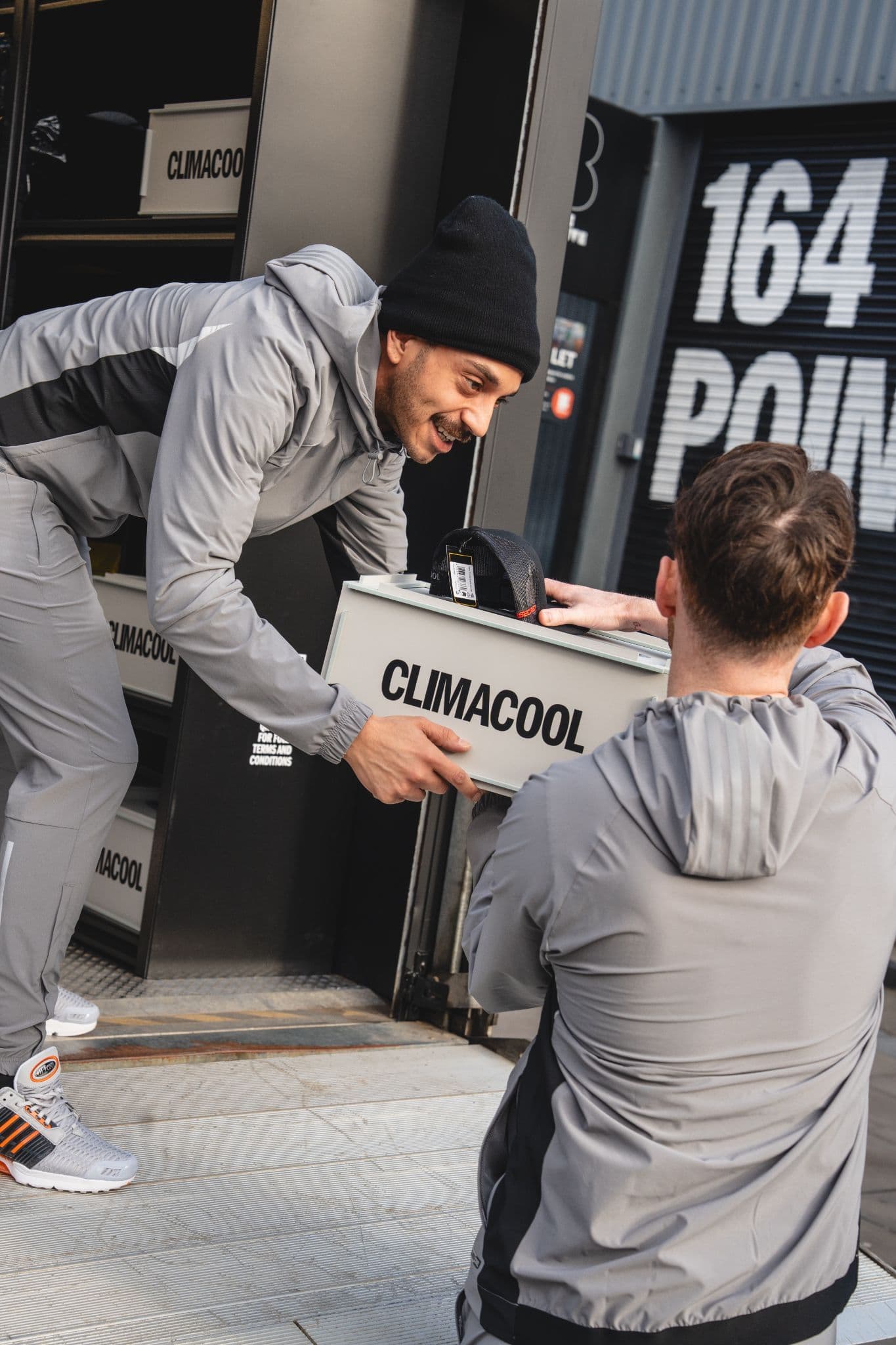 Two men in grey tracksuits unloading a branded Climacool box containing merchandise from a delivery vehicle.
