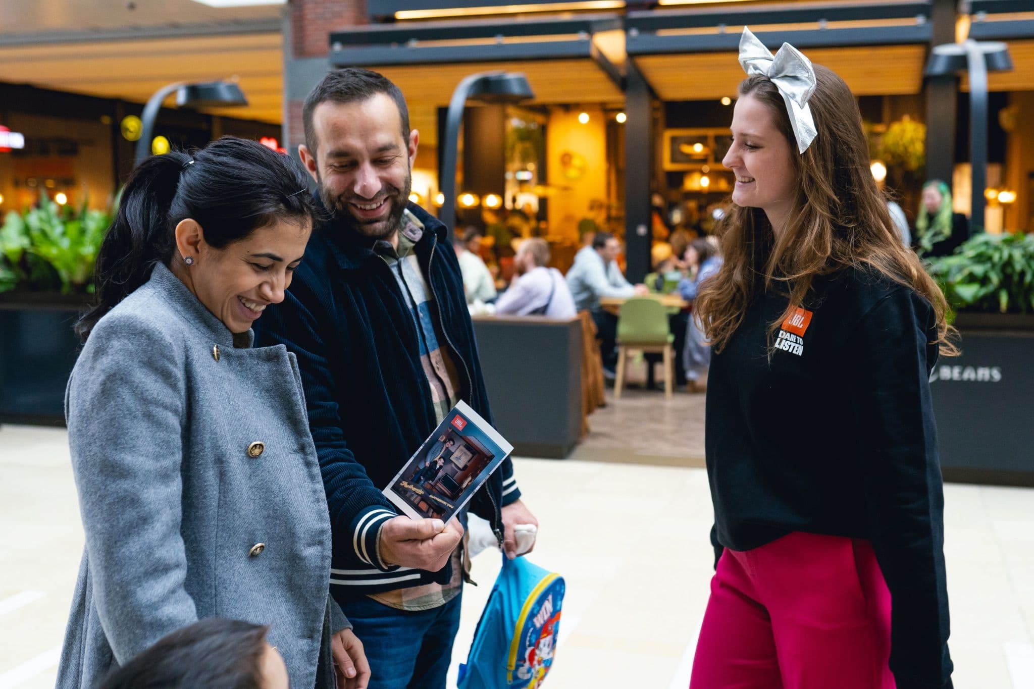 Smiling couple looking at a souvenir photo with a JBL staff member at a promotional stand in a shopping center.
