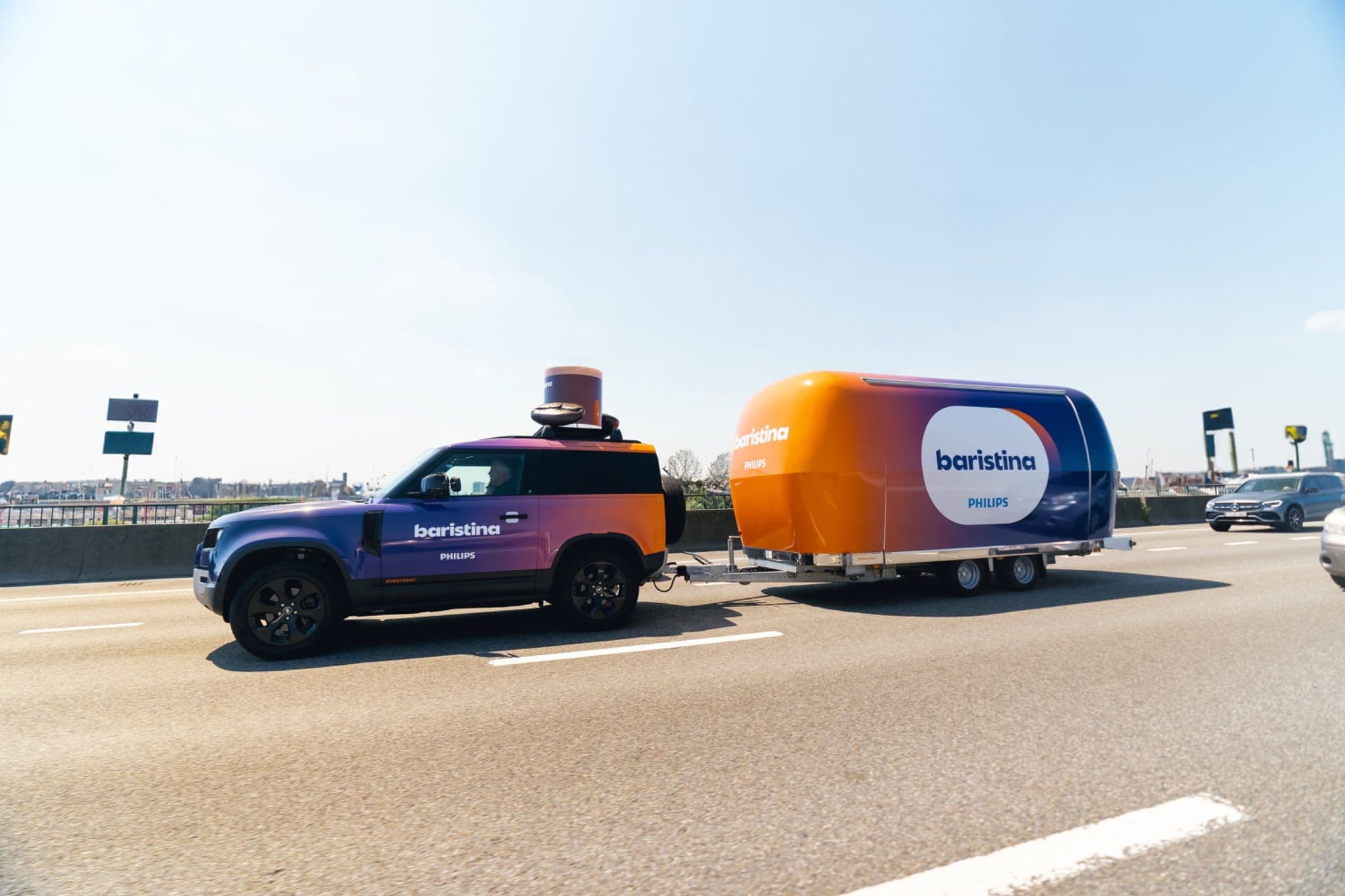Philips Baristina branded car and mobile coffee trailer wrapped in orange and purple gradients driving on a highway.