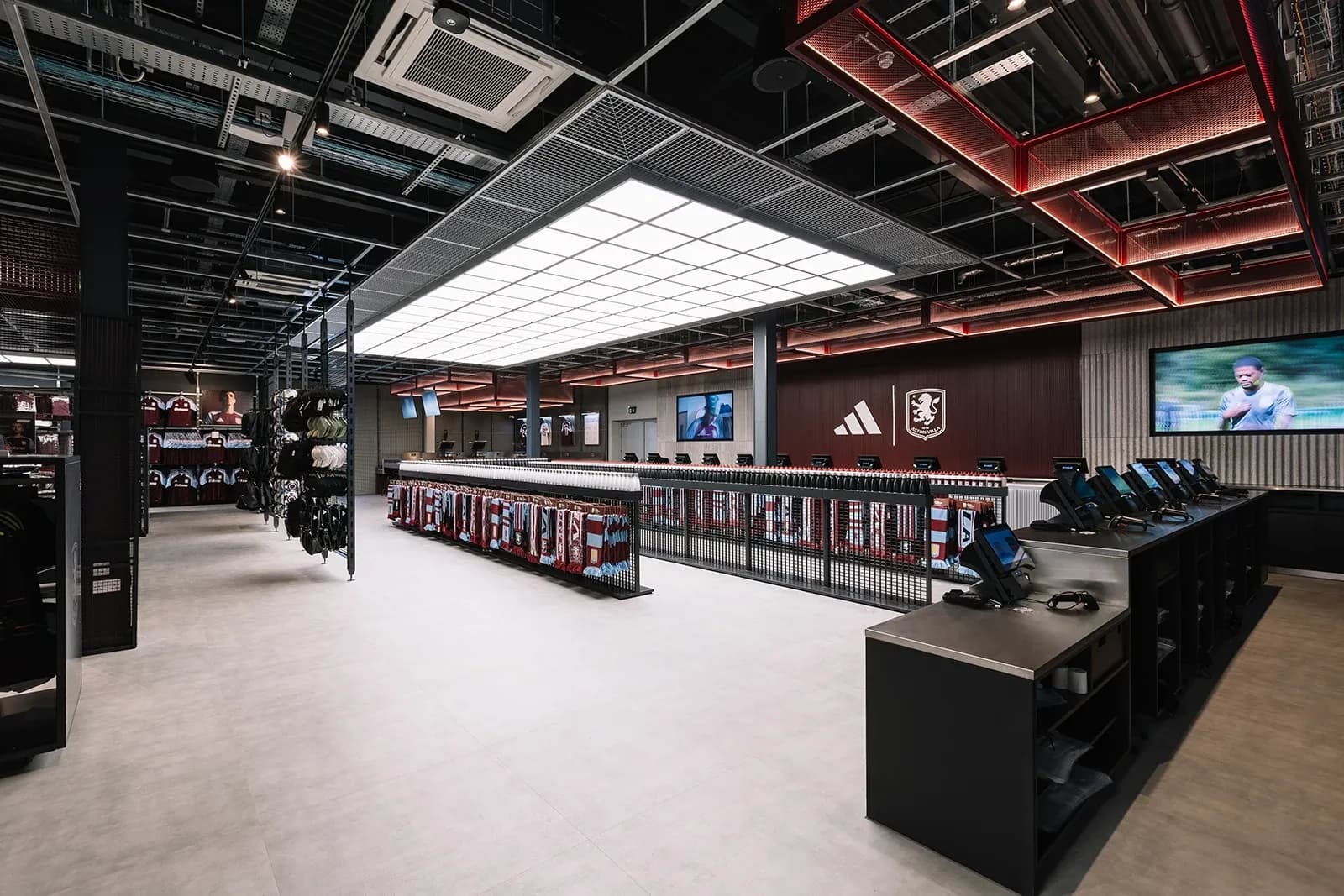 Wide view of the modern Aston Villa store checkout area with Adidas branding, digital screens, and industrial ceiling grid.