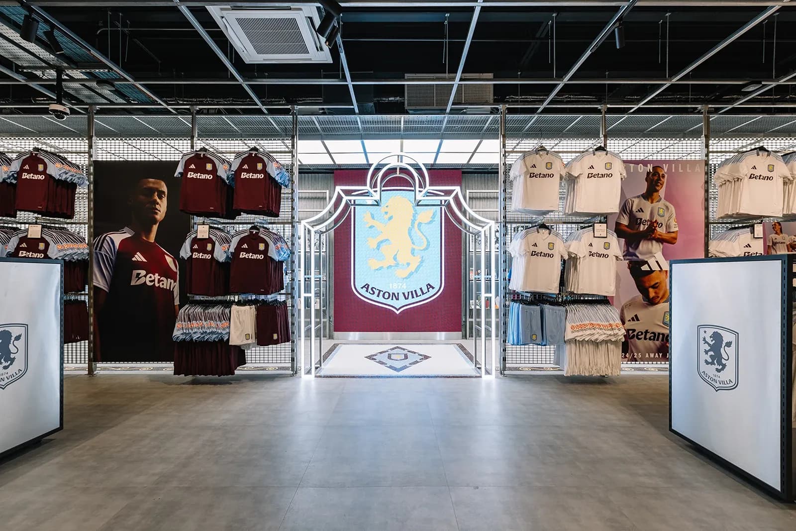 Illuminated neon Aston Villa crest archway surrounded by racks of white and maroon Adidas team jerseys.