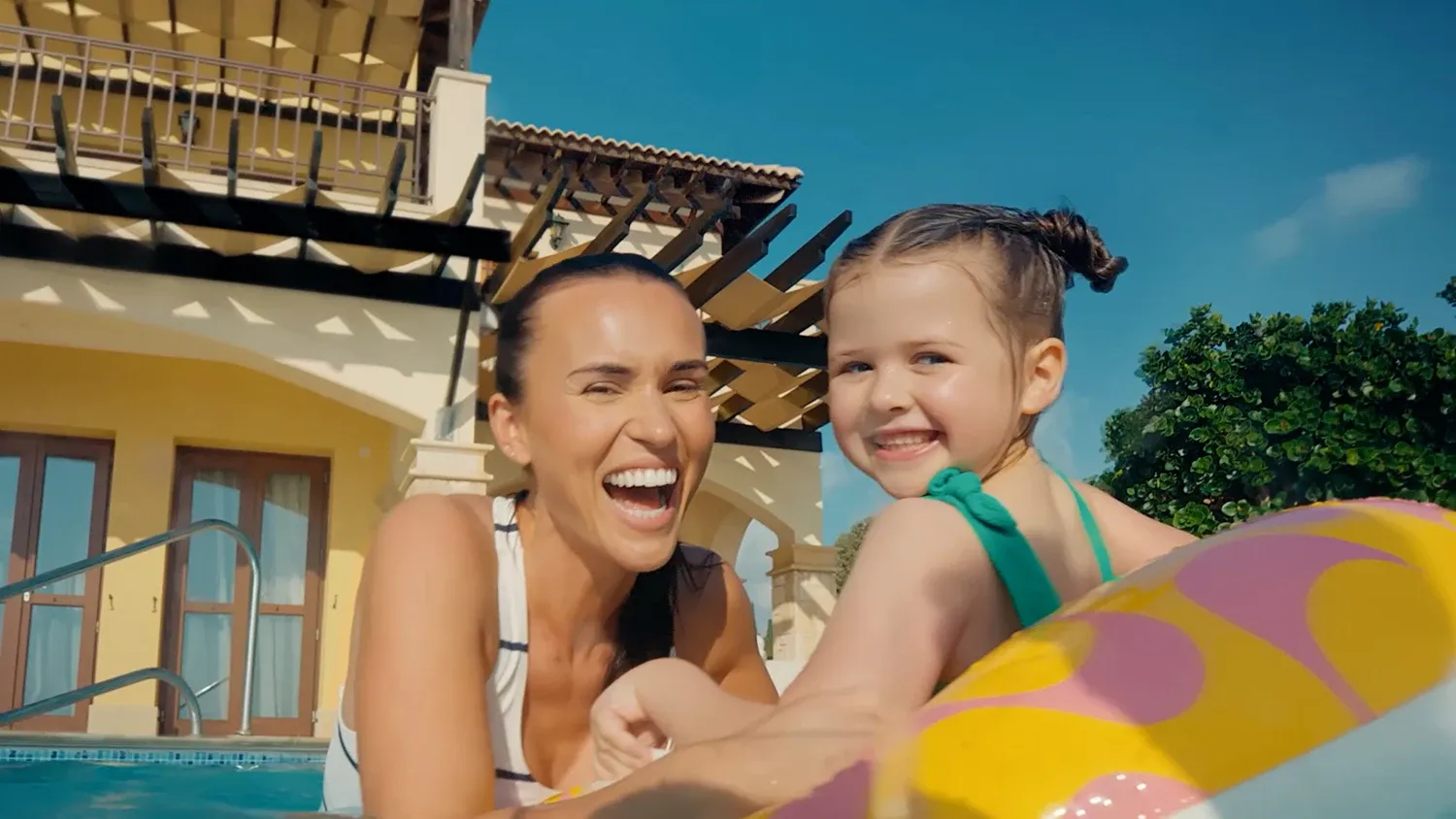 Smiling mother and daughter playing with a yellow inflatable ring in a pool outside a sunny holiday villa.